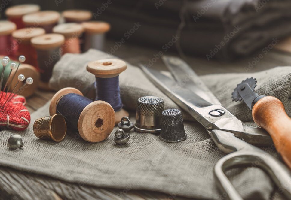 Retro sewing items: tailoring scissors, cutting knife, thimble, wooden thread spools, cushion for including pins, fabrics and sewing accessories.